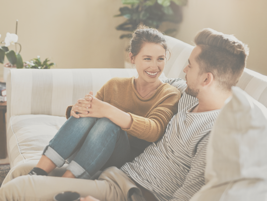 am woman and man sat smiling at each other in a partial embrace on a beige sofa